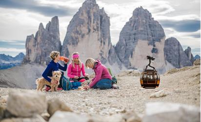 Three female hikers with a small dog are sitting on rocky terrain; the Three Peaks are in the background. A gondola is shown on the right side of the image.