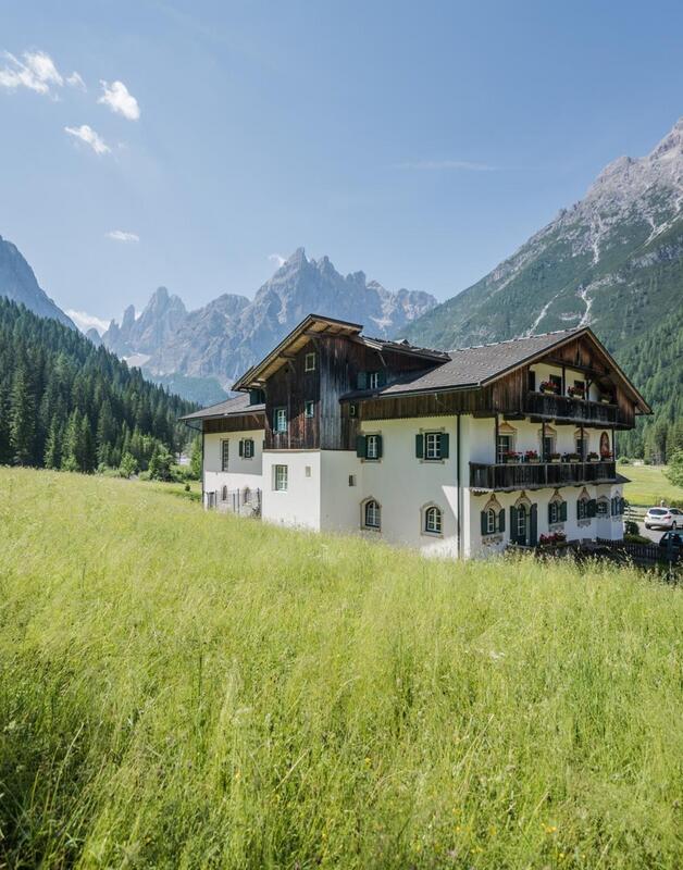 Edificio alberghiero tradizionale su un prato davanti a un bosco e al paesaggio montano delle Dolomiti.